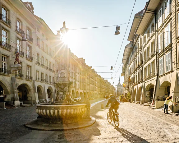 A cyclist rides down a sunlit cobblestone street, flanked by historic buildings and a decorative fountain, creating a vibrant city scene.