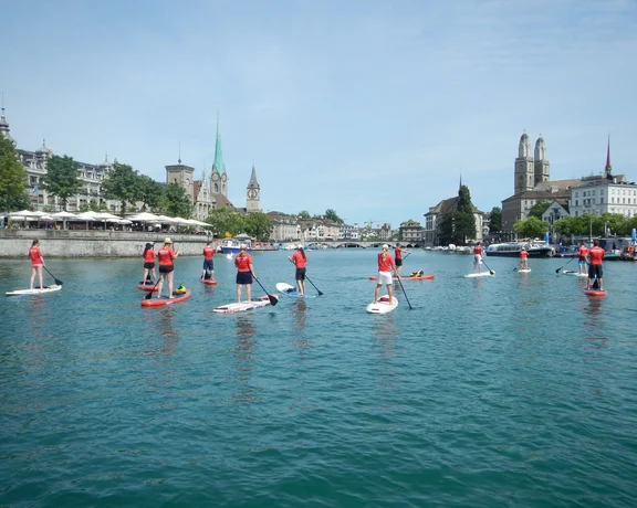 A group of paddleboarders in red shirts navigate the clear waters of Zurich, with historic buildings and a church steeple in the background.