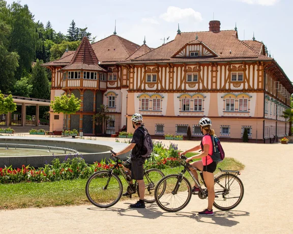 Two cyclists pause near a beautiful, historic building with a lively garden and fountain on a sunny day.