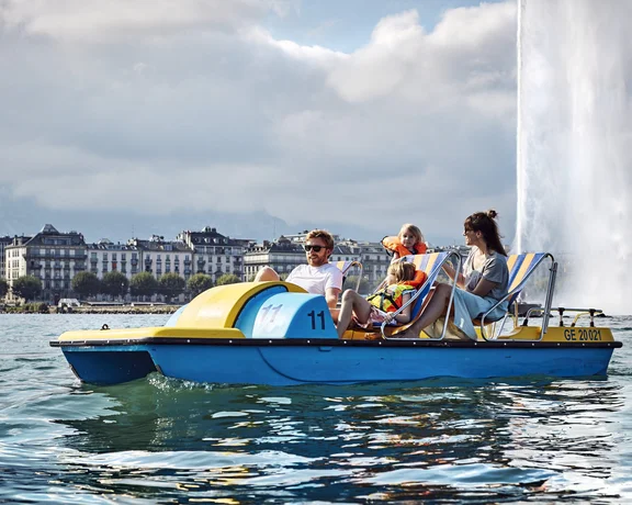 A family enjoys a peaceful pedal boat ride on the Geneva lake, with iconic city buildings and a fountain in the background.