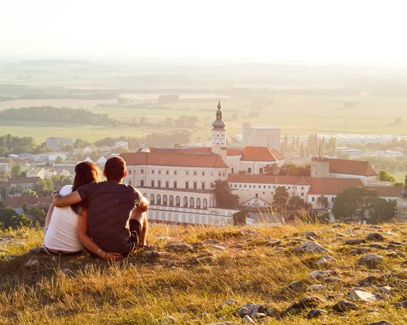 A couple sits on a grassy hill at sunset, overlooking the historic town of Mikulov in South Moravia. The view features the iconic Mikulov Castle with its red rooftops, Baroque-style architecture, and a central tower, surrounded by rolling vineyards and open countryside.