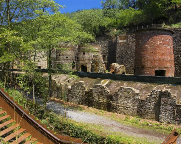 Ruins of an old industrial site, featuring stone walls and brick structures, surrounded by lush greenery and a gravel pathway.