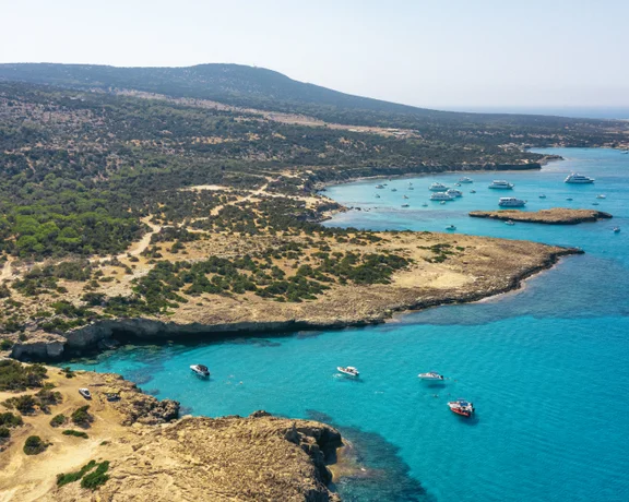 Aerial view of a serene coastal landscape with turquoise waters, boats anchored near rocky shores, and lush green hills in the background.