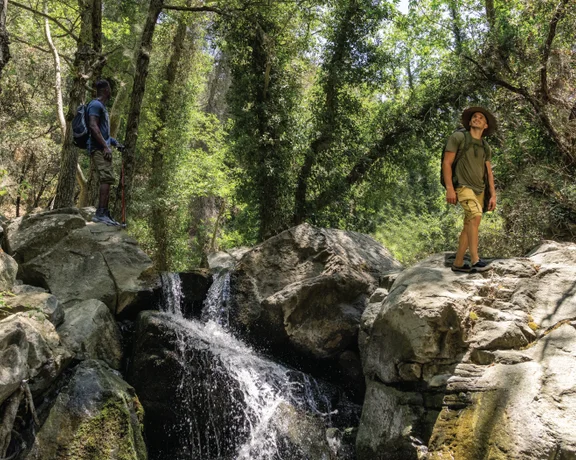 Two hikers stand atop large rocks beside a flowing waterfall in a lush, green forest, surrounded by trees and sunlight.