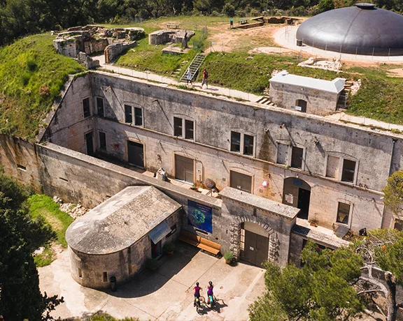 Aerial view of a historic fort with stone walls, grassy areas, and a round structure, surrounded by trees and visitors exploring.
