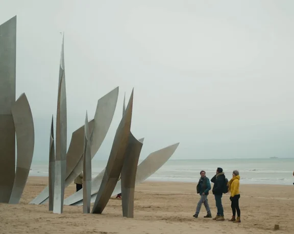 Three people admire a large, modern metallic sculpture on a sandy beach under a cloudy sky, with the ocean in the background.
