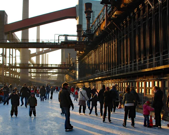 An industrial heritage site with crowds ice skating on a frozen path, framed by steel structures and towering chimneys.