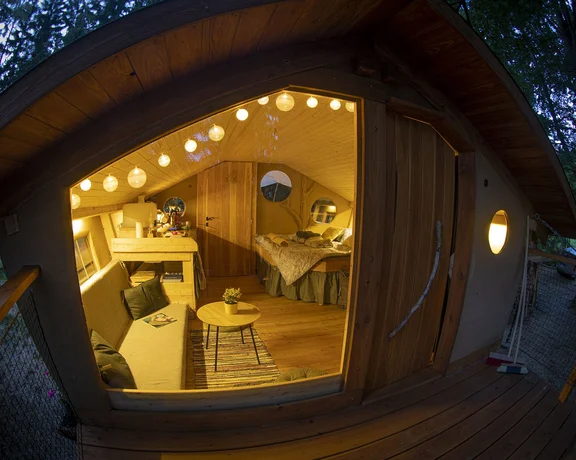 Cozy treehouse interior with wooden decor, soft lighting, a seating area, and a bed, framed by lush greenery outside.