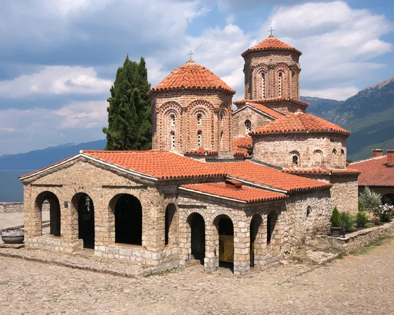 A historic Orthodox monastery with red-tiled roofs and stone arches, situated by a tranquil lakeside with mountains in the background.