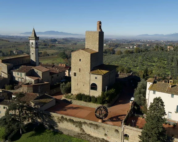 Aerial view of a medieval Italian village with stone buildings, a tall church tower, and terraced olive groves under a clear sky.