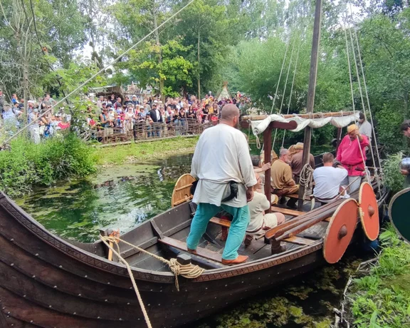 Traditional Viking boat with costumed participants on a riverbank, surrounded by spectators.