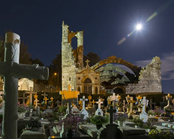 A moonlit cemetery featuring ornate crosses and a majestic ruined church, surrounded by lush greenery and colorful flowers.