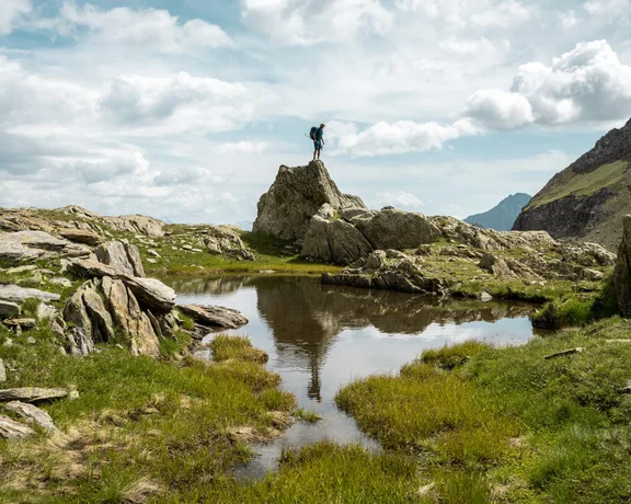 A hiker stands triumphantly on a rocky outcrop, reflecting in a serene pond surrounded by lush grass and mountains under a cloudy sky.