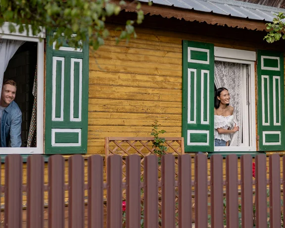 Two people look out from windows of a charming wooden house with green shutters, framed by a wooden fence and greenery.