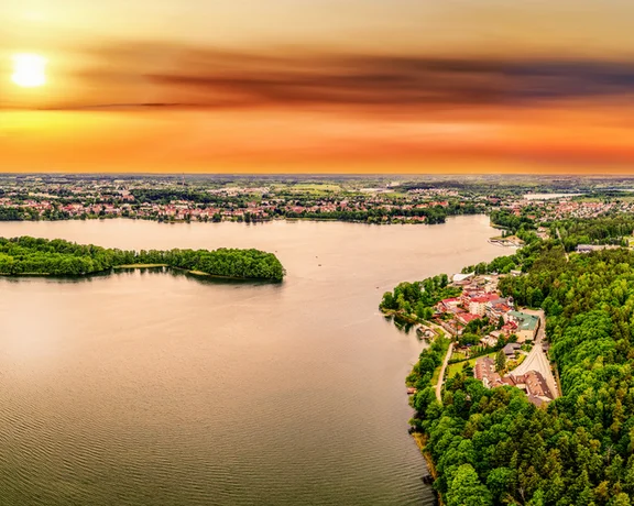 Aerial view of a shimmering lake surrounded by lush greenery and a quaint village, illuminated by a vibrant sunset sky.