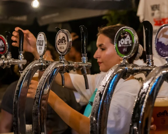 A hand reaches for a beer tap among several shiny taps, with various beer labels visible in a lively bar setting.