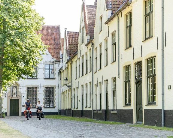 Two cyclists ride along a cobblestone path flanked by charming, historic buildings and a lush green tree in bright sunlight.