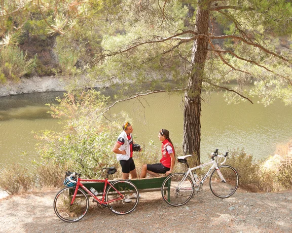 Two cyclists chat seated on a bench by a serene river, surrounded by trees and their bikes parked nearby.