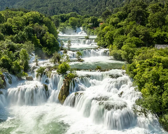 Aerial view of cascading waterfalls surrounded by lush greenery and dense forest under bright blue skies.