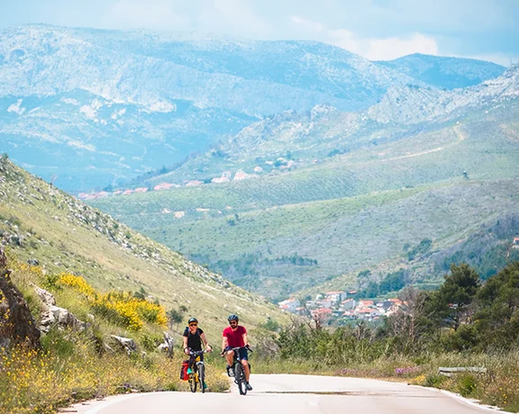 Two cyclists ride along a winding road, surrounded by lush hills and mountains under a partly cloudy sky.
