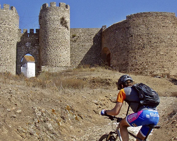 A cyclist approaches ancient stone fortifications on a dry hillside under a clear blue sky.