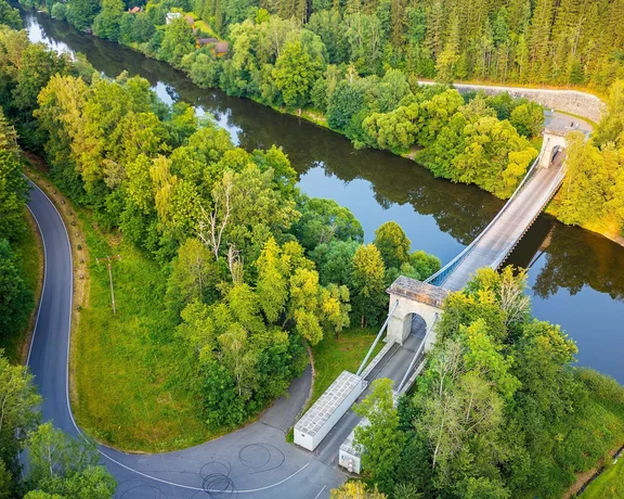 Aerial view of a curving river surrounded by dense greenery, featuring a bridge crossing the water and a winding road nearby.