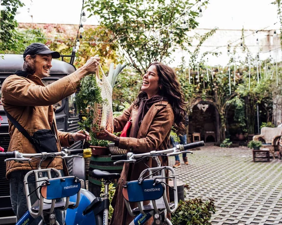 A couple exchanges fresh vegetables in a vibrant, green courtyard filled with plants and bicycles, creating a lively atmosphere.