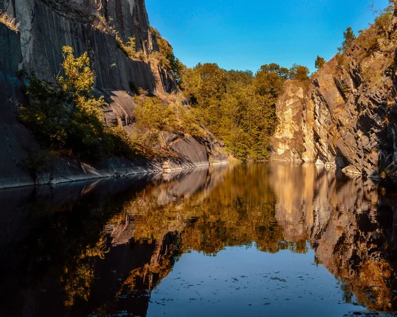 A tranquil quarry lake surrounded by steep, rocky cliffs and vibrant autumn foliage reflected in the calm water under a clear blue sky.