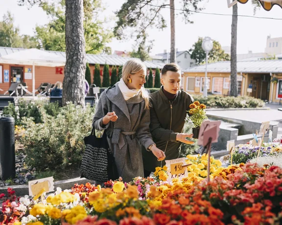 A woman and a man explore a vibrant flower market, surrounded by colorful blooms and sunny greenery.