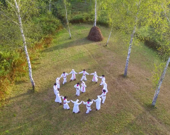Aerial view of dancers in traditional white costumes forming a circle in a clearing surrounded by birch trees during the Jurjevanje folklore festival.