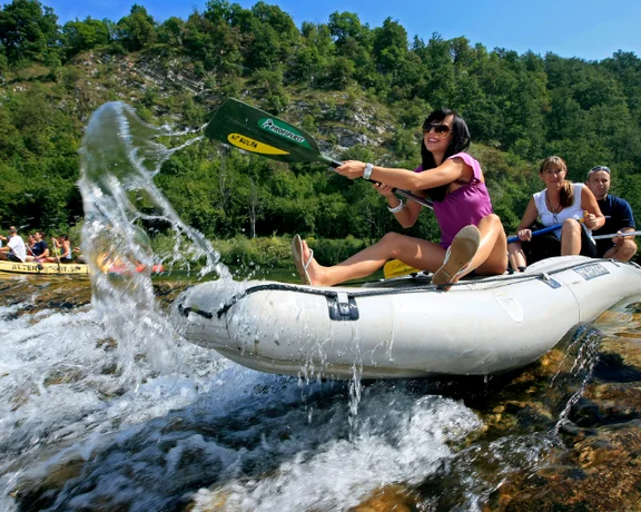 A group of people in a raft navigating through a rocky river with splashing water, surrounded by lush green hills under a clear sky.