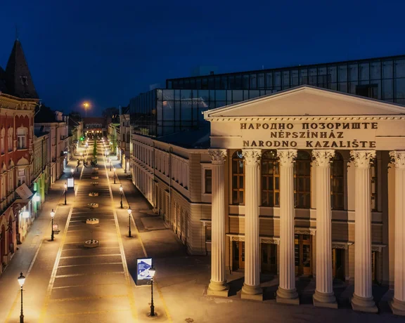 A beautifully illuminated street at night featuring the impressive facade of a theater with classical columns and charming buildings.