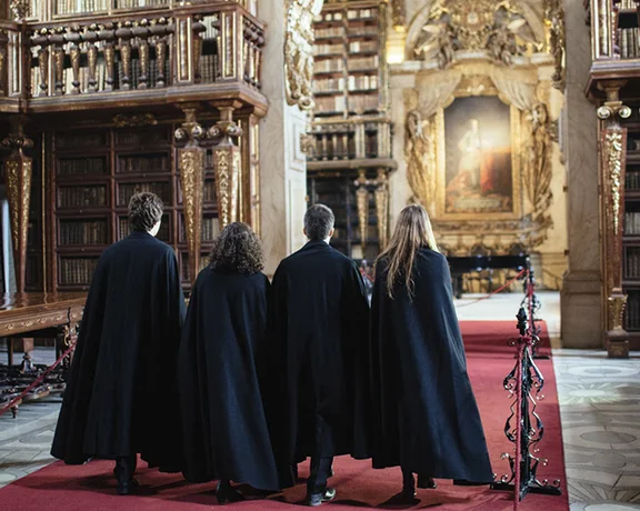 Four individuals in black cloaks walk through an ornate library, surrounded by bookshelves and historic artwork.