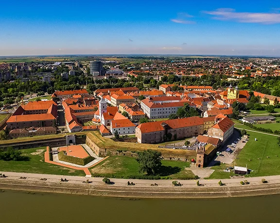 Aerial view of a historic fortress with red-roofed buildings, surrounded by greenery and a river, under a clear blue sky.