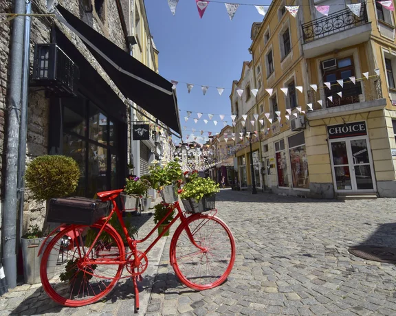 A cobblestone street in Plovdiv’s Kapana district decorated with colourful bunting, featuring a bright red bicycle adorned with flower baskets.