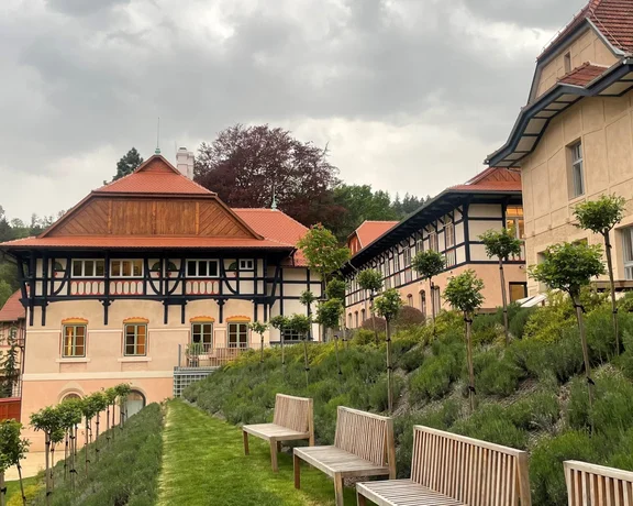 Historic buildings with wooden accents rise above manicured lawns, framed by lush greenery under a cloudy sky.