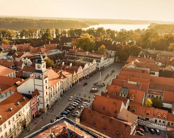 Aerial view of a charming town square with red-roofed buildings and a clock tower, overlooking a serene river and lush trees at sunset.