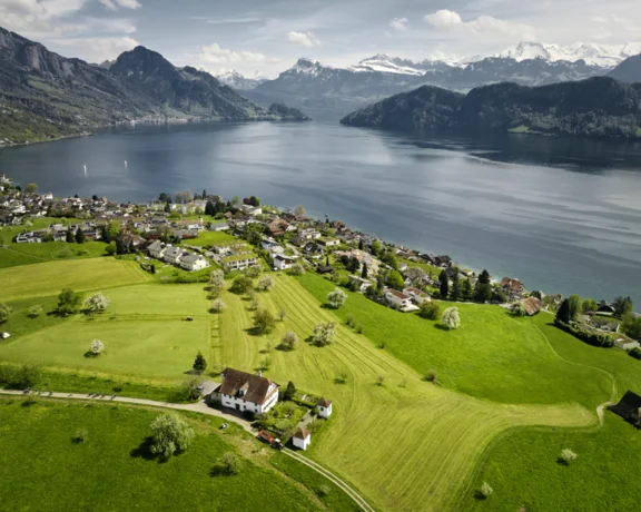 Aerial view of a picturesque lakeside village surrounded by lush green fields and mountain scenery, with snow-capped peaks in the background.
