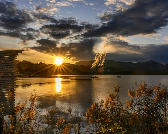 Velenje Lake in Slovenia, reflecting the golden sunset with a view of mountains and steam rising in the distance.