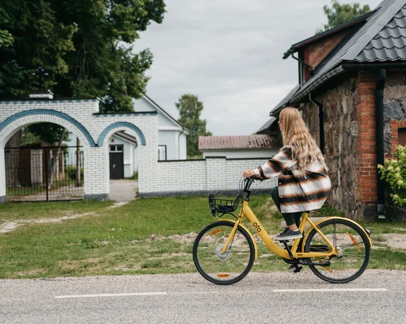 Young woman riding a yellow rental bike past a rural house and white brick archway on a cloudy day.