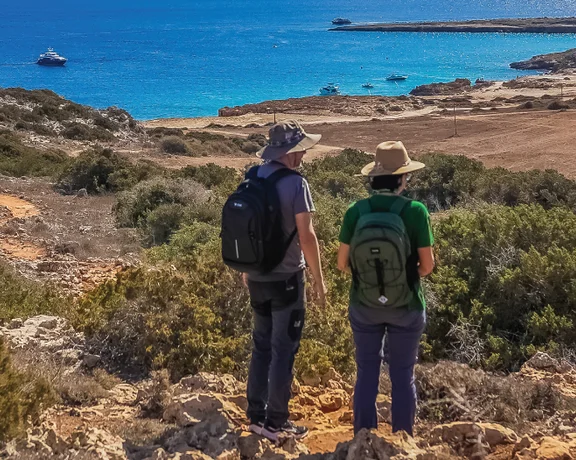 Two hikers with backpacks admire the rocky coastline and bright blue sea from a trail in the Akamas Peninsula.