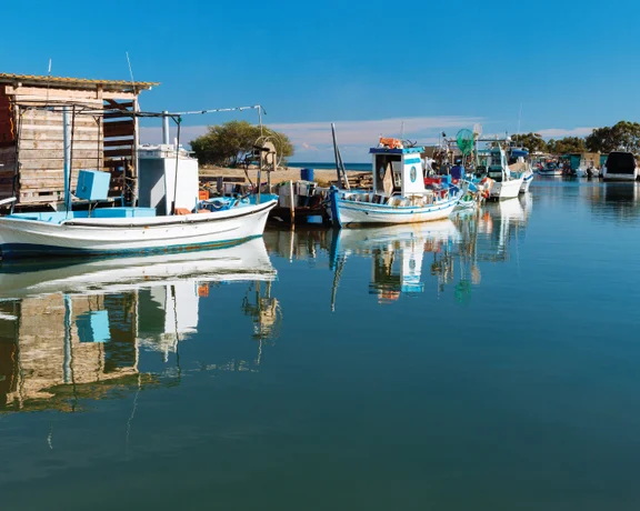 Traditional fishing boats docked along the calm waters of Liopetri River, reflecting on the surface under a clear blue sky.
