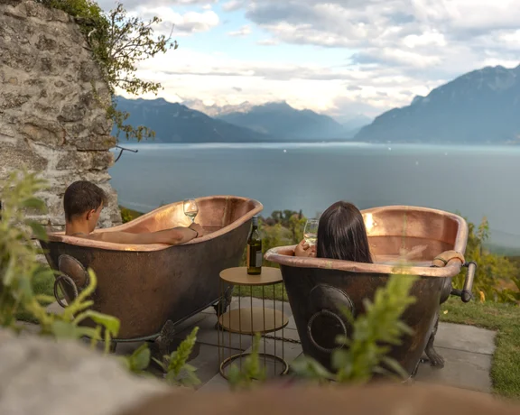 Couple relaxing in outdoor copper bathtubs with wine, overlooking Lake Geneva in Switzerland.