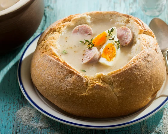 Polish traditional soup with egg and sausage served inside a bread bowl, placed on a rustic blue wooden table.