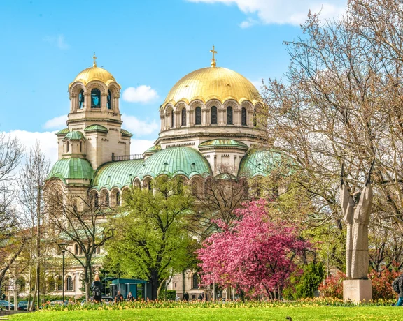 Alexander Nevsky Cathedral in Bulgaria, partially hidden by blooming pink and green trees, under a clear blue sky.