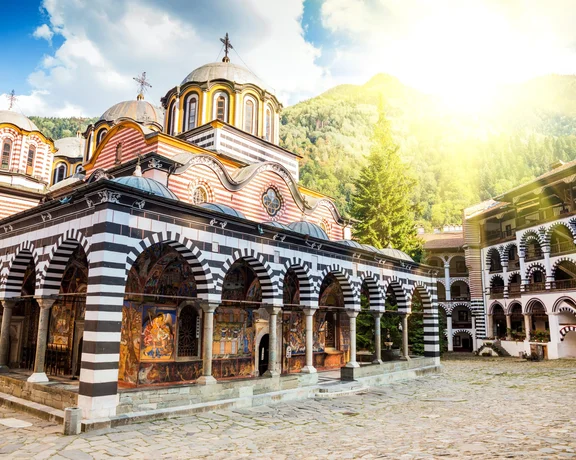 Rila Monastery in Bulgaria with its distinctive striped arches, set against a backdrop of mountains at sunrise.