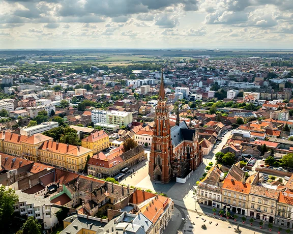 Aerial view of the city of Osijek in eastern Croatia, featuring the prominent neo-Gothic Co-Cathedral of St. Peter and St. Paul with its tall spire rising above red-tiled rooftops. The cityscape extends into the distance with a mix of historical and modern buildings, under a partly cloudy sky.