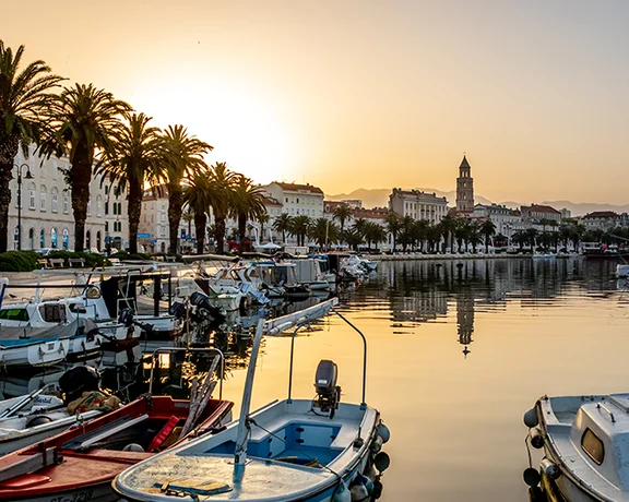 Sunrise over Split, Croatia, with boats moored in the calm harbor and historic buildings silhouetted against the glowing sky.