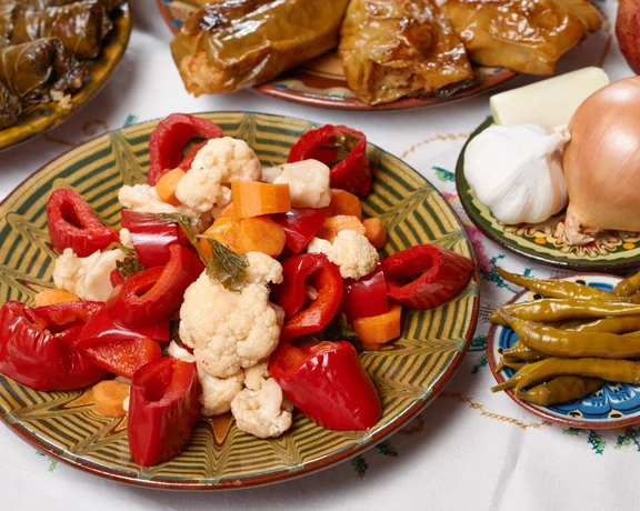 Traditional Bulgarian pickled vegetables with peppers, carrots, cauliflower, and herbs served on a ceramic plate alongside garlic and onions.