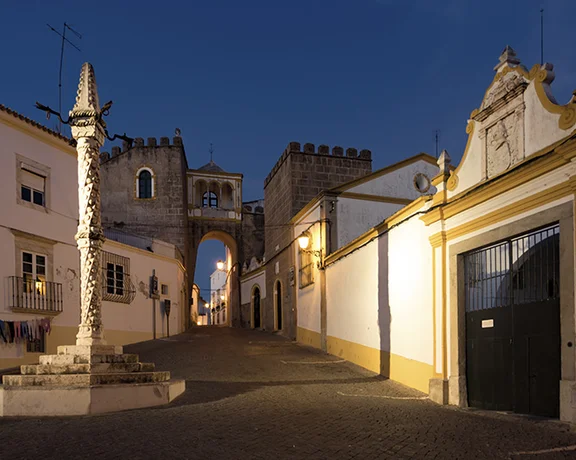Narrow street in Elvas at night, lined with historic buildings, illuminated by warm streetlights, with a sculpture prominently displayed in the center.